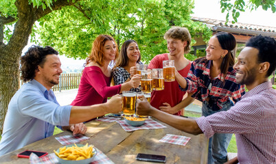 multiethic group of people enjoying beer together outdoor sitting on a garden table. diverse happy friends having fun making a toast drinking alcohol in summer. social, holidays and youth concept