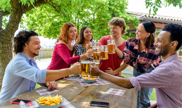 Multiethic Group Of People Enjoying Beer Together Outdoor Sitting On A Garden Table. Diverse Happy Friends Having Fun Making A Toast Drinking Alcohol In Summer. Social, Holidays And Youth Concept