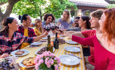 multiethic big group of people enjoying barbecue dinner together outdoors sitting on garden table eating and toasting wine. large diverse happy friends having a summer evening picnic. focus on glasses
