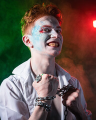 Aggressive psycho with blood on his face and chain in his hands against the background of smoke, illuminated by red and green light
