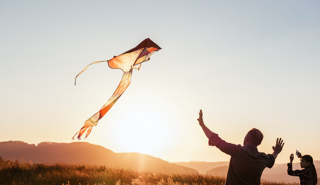 Sunset sun backlit silhouettes of a daughter with father while they trying to launch a colorful kite in the mountain fields. Warm family moments or outdoor time spending concept image.