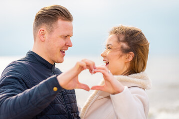 Two young couple is making heart, looking at each other and smiling while standing at the beach.