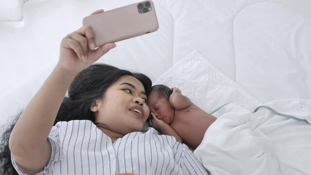 Mother and her newborn baby making a selfie or video call to family on the bed white in home. Mixed race black boy Ethnicity Thai-Nigeria. New generation Technology Communication shot in slow motion