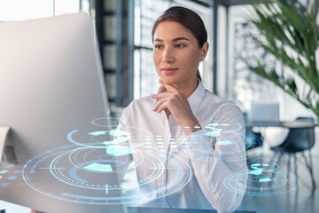 Attractive businesswoman in white shirt at workplace working with laptop to optimize development by implying new technologies in business process. Hi tech hologram over office background
