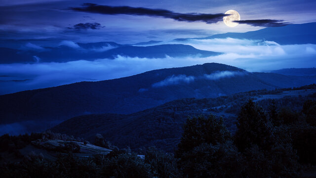 rural landscape at night. mist in the distant valley in full moon light. silhouette of trees and hills