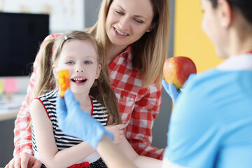 Doctor holding jar of capsules and red apple in front of child and mother in clinic