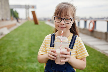 Waist up portrait of smiling girl with down syndrome holding milkshake outdoors and looking at...