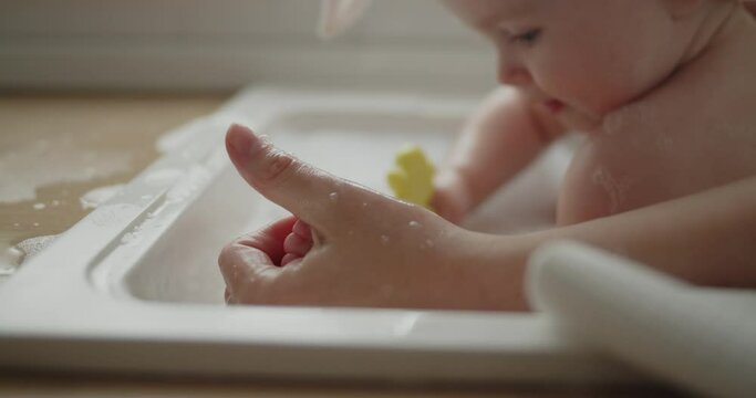 Mother and daughter spend time at home together. Young mom helps her adorable little child to take a bath in a kitchen sink. Adorable girl have fun in the water and foam.