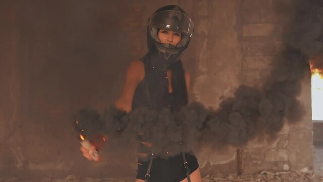 young female in motorbike helmet holding colored smoke bomb slow motion on the background abandoned building