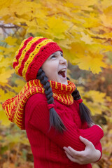 A girl with dark hair in a red cap and a large scarf shouts, is capricious looking to the side in an autumn forest or park against a background of yellow leaves on maple branches