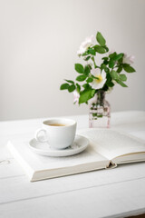 Eine weiße Tasse Kaffee auf einem offenen Buch und frische Blumen in einer Vase. Stillleben,...