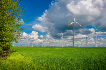 Panoramic view over beautiful farm landscape with green wheat field, green forest and family of wind turbines to produce green energy in Germany at Spring, blue dramatic rainy sky and sunny day.