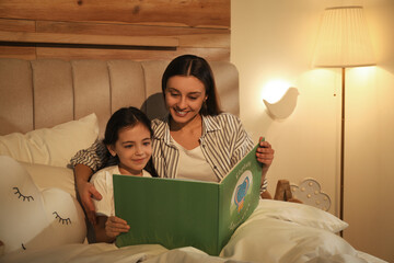 Little girl with mother reading book in bedroom lit by night lamp