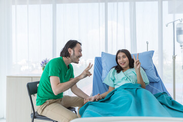 Obraz premium Happy young Asian couple with casual colthes is touching and holding wife patient's hands while showing v-sign to encourage and make patient feels relax and smile, on hospital bed in hospital.