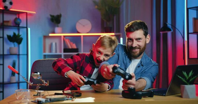 Leisure family concept where attractive happy smiling bearded dad and teen son enjoying videogames and celebrating victory with raised hands ,front view