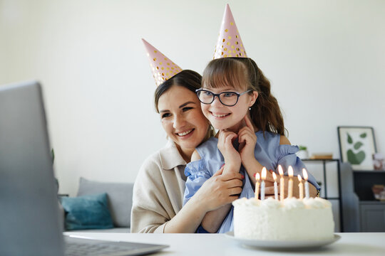 Portrait of happy mother and daughter with disability celebrating birthday online and blowing candles by laptop