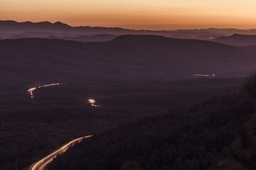 Looking down from a forest mountain ledge at the roadway trailing through a pine forest at night shows light trails in bright red and orange from vehicles driving the highways below