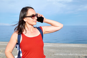 Beautiful woman in sun glasses and red top talking on cell phone while walking on beach with sea in background. Communication and connectivity concept