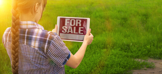 Girl holds tablet with for sale text, background of green field, plot of land for house construction, banner photo
