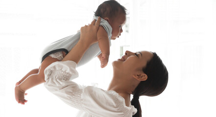 Portrait of enjoy happy love family asian mother playing with adorable little asian baby.Mom touching with cute son moments good time in a white bedroom.Love of family concept