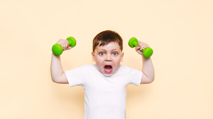 A little cute kid boy in a white t-shirt with an open mouth demonstrates his strength by raising his hands with dumbbells isolated on light color beige background. A sporty child does exercises