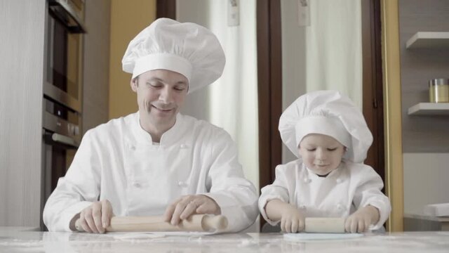 Dad and son rolling out blue sugar paste in their home kitchen. Hands of father and son using rolling pin. Father teaching son cooking a cake at home.