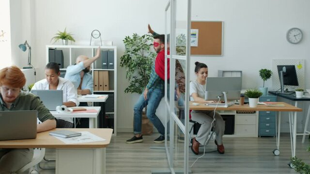 Funny young man in super hero costume is running in office doing high-five with colleagues wearing red cape and mask. Superman and workplace concept.