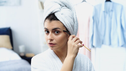 focused young woman with head wrapped in towel styling eyebrow with cosmetic brush in bedroom
