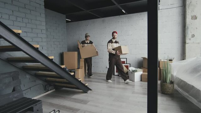 Slowmo tracking of male movers in uniform carrying cardboard boxes into empty house or apartment