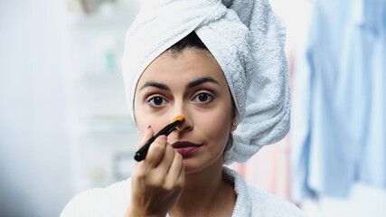 young woman with head wrapped in towel applying foundation on nose with cosmetic brush in bedroom