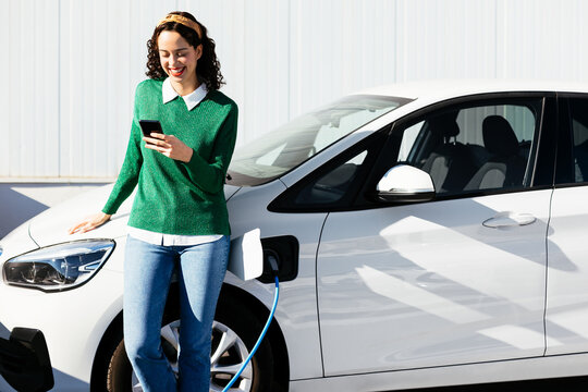 Optimistic woman browsing smartphone while recharging electric vehicle