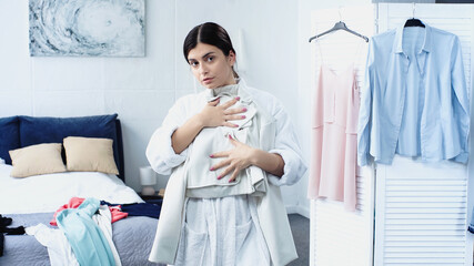 young woman in bathrobe holding jacket near bed with clothes in bedroom