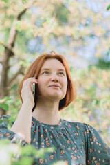 Young redhaired middle age woman wear casual dress holding smartphone in raised hand and focused on listening. Interesting conversation in spring park.