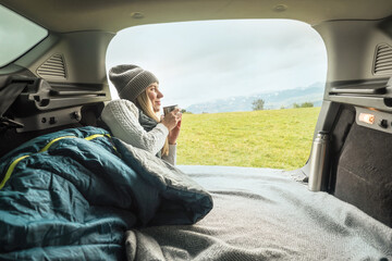 Girl resting in her car. Woman hiker, hiking backpacker traveler camper in sleeping bag, drinking hot tea and relaxing on top of mountain. Health care, authenticity, sense of balance and calmness.
 - obrazy, fototapety, plakaty