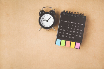 black analog alarm clock and opened calendar with multi color paper tag on grunge brown paper...