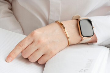 Feminine hands wera smartwatch with black screen and golden bracelet lying on day planner with white pages.