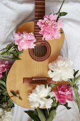 An acoustic wooden guitar lies on a white sheet surrounded by pink and white peonies.