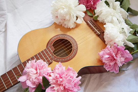 An acoustic wooden guitar lies on a white sheet surrounded by pink and white peonies.