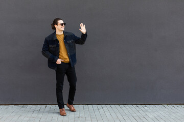 Young stylish man wearing suit and sunglasses standing outdoors on the city street waving to friend looking aside smiling suprised