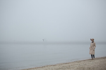 Fototapeta na wymiar Foggy morning at sea. Woman in hat and coat walking near autumn foggy sea