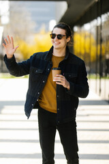 Young positive casual man with coffee to go happily waving hand hello on city street