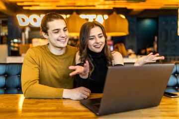 Handsome man and pretty woman making video call to their friend using free wi-fi on generic laptop pc, having meal, sitting at table in modern cafe interior