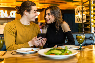 Cheerful young couple browsing internet on smartphone during romantic dinner at cozy cafe. Handsome boyfriend and girlfriend watching video together on cellphone while having lunch