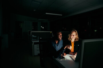 Two diverse happy colleagues working on laptop until night in dark office