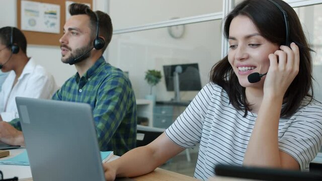 Young lady call center operator is talking to customer and smiling using laptop at work while colleagues male and female are busy in background
