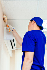 A painter in a cap and gloves paints the wall with a paint roller. Portrait of a young worker in uniform. Professional repair of premises. Mockup