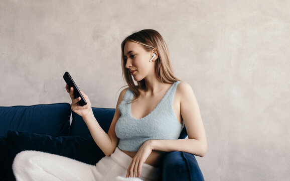 Relaxed young lady in true wireless earphones using smartphone on sofa