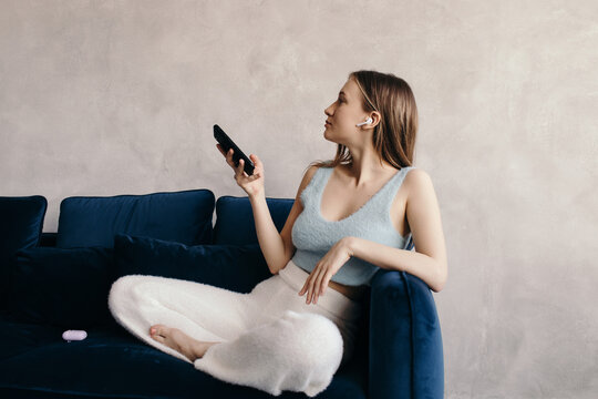 Relaxed young lady in true wireless earphones using smartphone on sofa