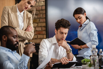 thoughtful businessman looking at notebook near multiethnic colleagues