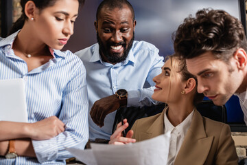 cheerful african american businessman looking at documents near blurred colleagues